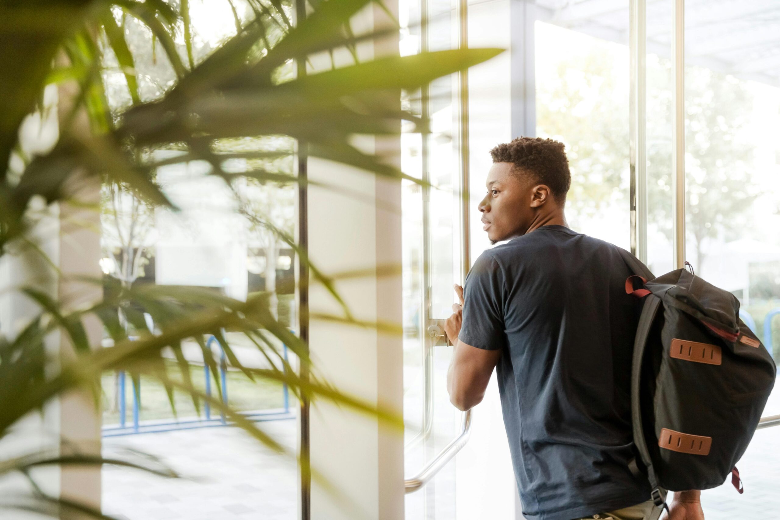 A Nigerian male student looking outside window carrying black and brown backpack while holding his hand on window