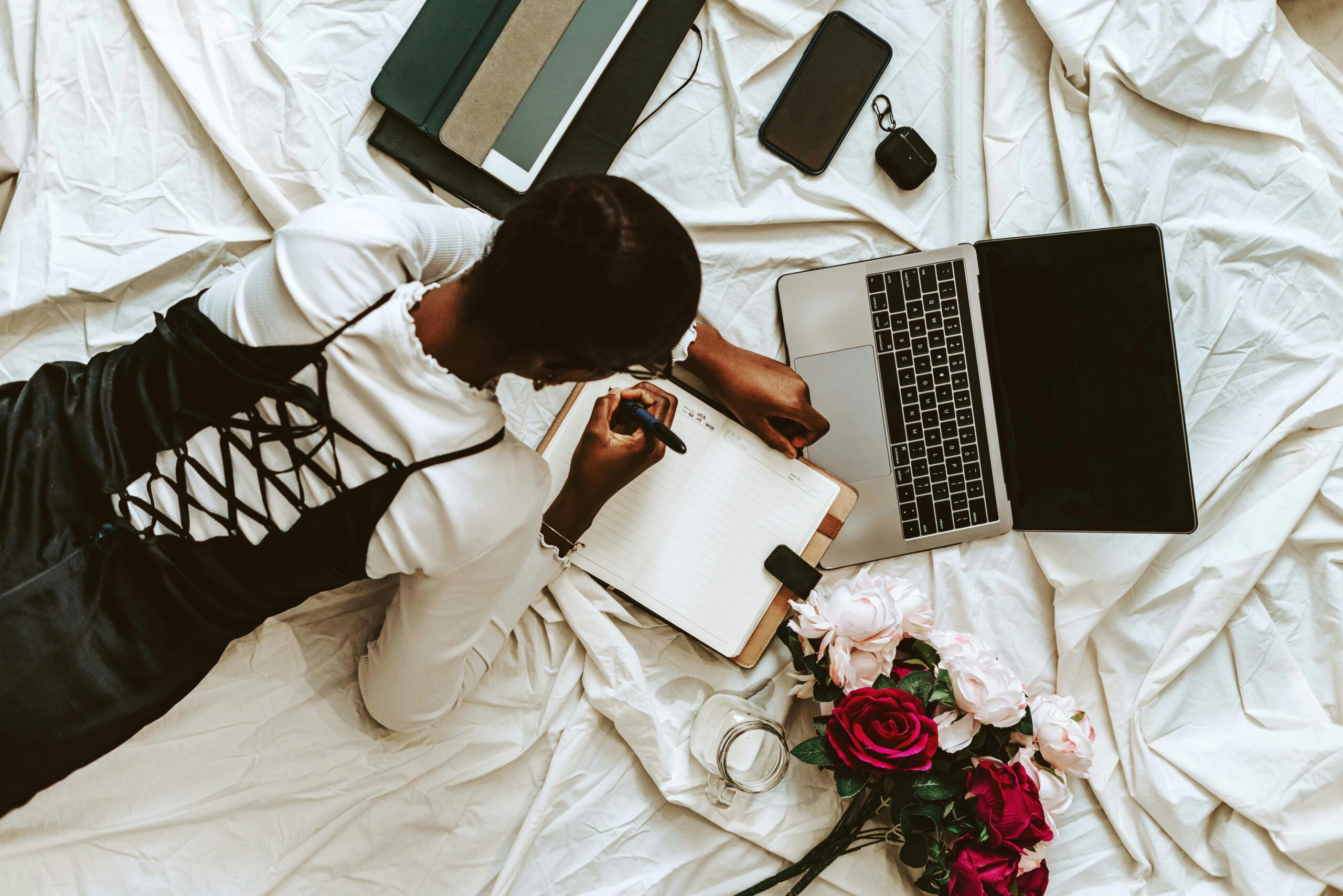 A Nigerian female student laying on a bed with a laptop, books and flowers