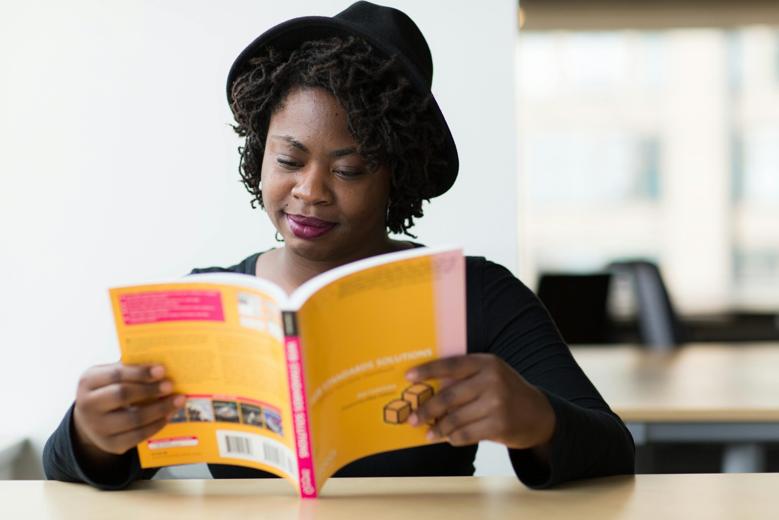 A Nigerian female student in long-sleeved shirt reading a yellow covered book while sitting down